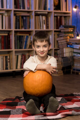 Fototapeta premium A child sitting on a checkered blanket, holding a large pumpkin. Cozy bookshelf background filled with books and string lights. Ideal for autumn and educational themes.
