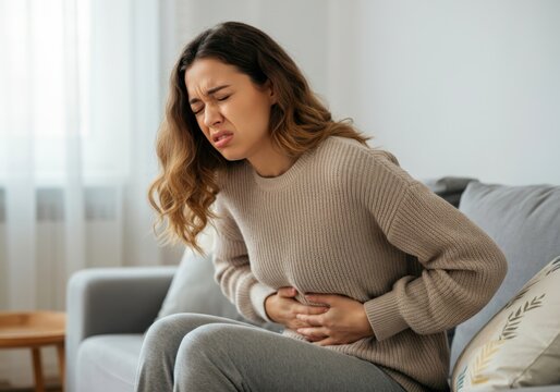 Woman experiencing severe abdominal pain, holding her belly with discomfort and closed eyes, sitting on a sofa in the living room