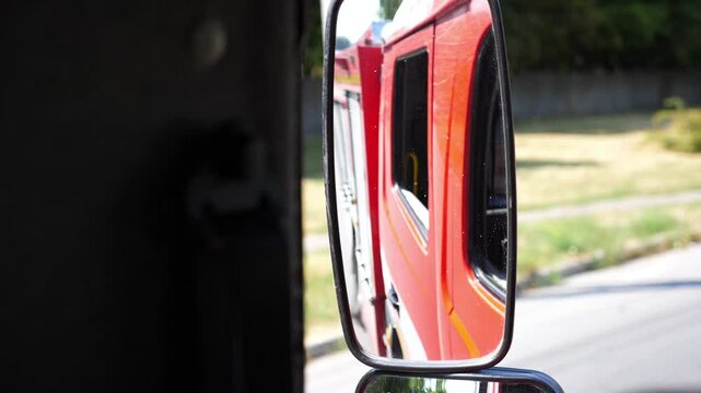 View to side mirror of fire engine riding to an emergency. Red big truck driving to place of a fire on sunny summer day. Concept of saving lives and heroic profession. Close up Slow motion