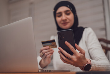 A young happy Muslim woman using mobile phone and holding credit card checking balance account or...