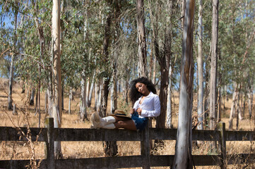 Fototapeta premium woman, young, beautiful, Latin, brunette with curly hair dressed in white shirt, denim skirt, white boots and hat, sitting with her legs stretched out on the forest fence. The woman rests.