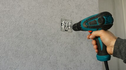 An employee repairing or servicing an electrical outlet or switch using an electric screwdriver against a background of gray wallpaper