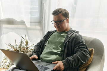 A young man with Down syndrome enjoys browsing on his laptop in a bright, comfortable living room.