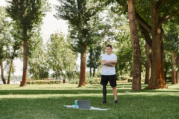 Tattooed man with artificial leg working out in green park, engaging in an online fitness lesson