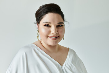 A stunning plus size bride radiates joy, adorned in a white gown and sparkling earrings on a serene grey background.