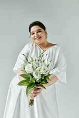 A radiant plus size bride beams as she cradles a bouquet of flowers against a soft, grey backdrop.