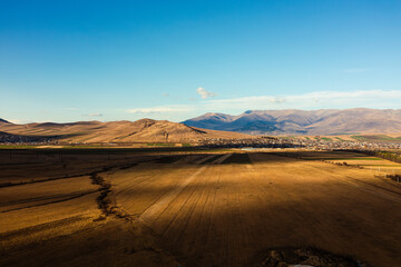 A vast landscape showcasing golden fields stretching into the horizon, bordered by a serene town and majestic mountains under a crisp, clear blue sky.