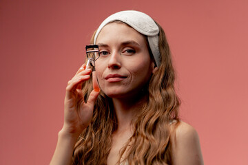 Fototapeta premium close up studio beauty shoot on pink background beautiful girl in white underwear and a sleeping mask on her head standing with eyelash curlers beauty salon