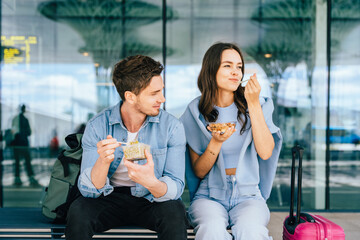 Young adults enjoying snacks while waiting at airport terminal. Two young adult friends, a Caucasian man and woman, eating snacks while sitting on the bench at airport terminal, outdoor.