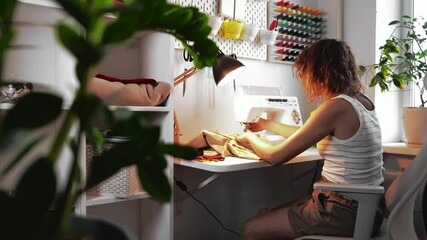 Young woman sews custom-made clothes, selects fabric to create unique, custom-made items. Seamstress works at table in comfortable chair in home workspace. Concept of custom tailoring, customer focus