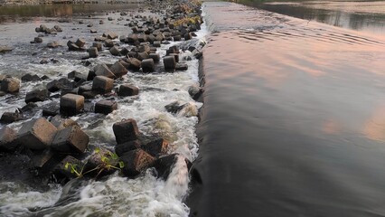 A river cascades over a concrete barrier, creating textured waves as it flows past angular rocks....