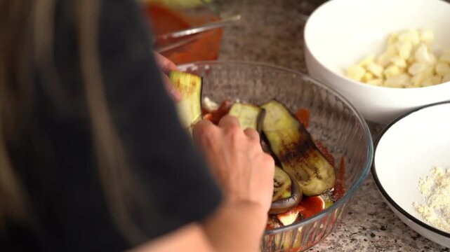 A young woman is putting roasted and sliced ​​eggplants on a plate to prepare the southern classic parmigiana in the kitchen. The concept of home cooking, healthy eating, fresh vegetables