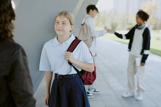 Smiling blonde Caucasian little girl dressed in school uniform with red wine backpack chatting with unrecognizable friend - Powered by Adobe