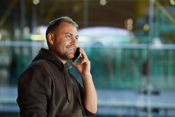 A man in casual clothes having a phone conversation on his smartphone, set against a modern glass building background.
