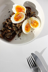 Toast with Soft Boiled Eggs, Fried Mushrooms with Onion, Garnished with Sesame Seeds and Black Pepper. Breakfast Served on White Table with Fork and Knife.