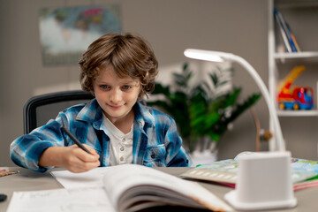 in the children's room a schoolboy in white sweater and a blue checkered shirt at a desk doing homework on his studies writing in a notebook