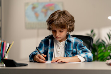 close up in the children's room a schoolboy in white sweater and a blue checkered shirt at a desk doing homework on his studies writing in a notebook