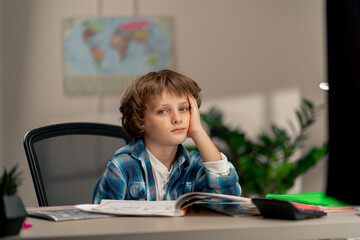 in the children's room a schoolboy in a white sweater and a blue checkered shirt at a desk doing...