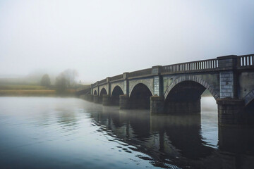 Fototapeta premium a fog-covered bridge over a river, with the water reflecting the misty environment, adding depth and atmosphere to the scene.