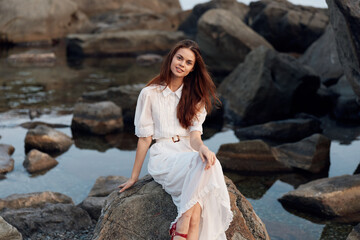 Serene summer seaside gorgeous woman in white dress relaxing on rocks by the ocean on a sunny day