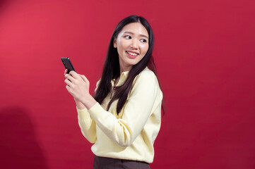 Naklejka premium Asian woman smiling while holding a smartphone with both hands, wearing a yellow sweater against a red background. The scene suggests casual engagement with technology or communication.