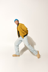 A young man wearing a yellow jacket and blue beanie poses dynamically in a stylish outfit.