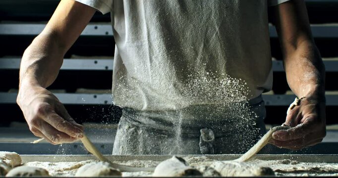 Super slow motion close up of professional artisan baker chef sprinkles flour on raw loaf of dough while making homemade bread, pasta or pizza on rustic wooden table in traditional bakery kitchen.
