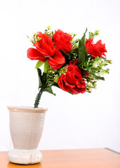 A bouquet of red flowers with green leaves and white filler flowers in a cream-colored vase with embossed designs, placed on a wooden surface against a white background.