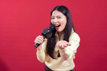 Obraz premium Asian woman smiling and singing into a microphone, wearing a yellow sweater with a red background. She appears enthusiastic and expressive, holding the microphone