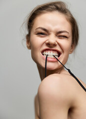 Cheerful young woman playfully biting sunglasses straps, expressing joy with a playful smirk. Light gray background emphasizes her vibrant energy and spontaneity.