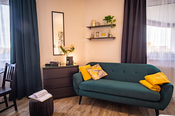 Green sofa with yellow pillows and a blanket in a hotel style room. Black curtains. 