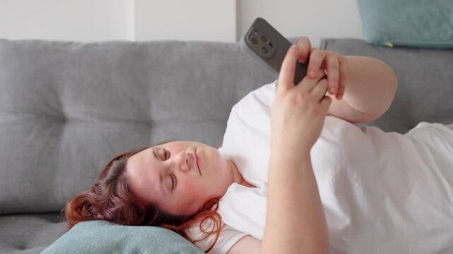 Lazy overweighted woman using her phone while laying on sofa. She is text messaging or writing on social media.