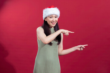 Asian woman wearing a Santa hat and a sleeveless green ribbed dress is pointing to the side with both hands, smiling cheerfully, against a solid red background. Her festive appearance and joyful