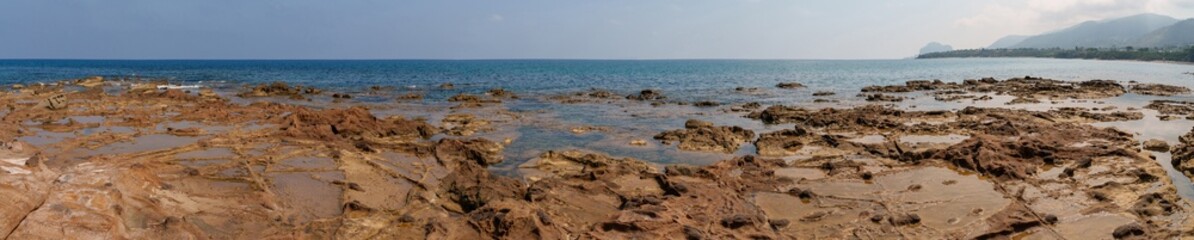 A panoramic view capturing a rocky coastline, calm sea, and distant hills under a clear sky. The expansive scene highlights the natural beauty and rugged terrain of the coastal landscape