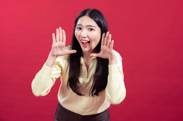 Fototapeta premium Asian woman posing playfully with hands framing her face, smiling widely, wearing a yellow sweater against a red background. Her expression is lively and engaging, showing enthusiasm and joy