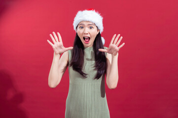 An Asian woman is joyfully raising both hands with an excited expression. She is wearing a Santa hat and a sleeveless green ribbed dress, standing in front of a solid red background