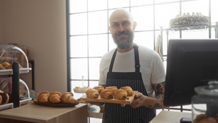 Handsome hispanic man with beard and tattoos holding pastries in bakery shop with indoor lighting