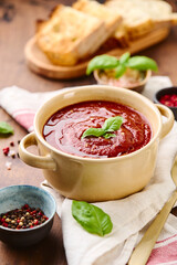 Tomato puree soup garnished with basil leaves and spices in ceramic bowl with toasted bread slices on wooden table