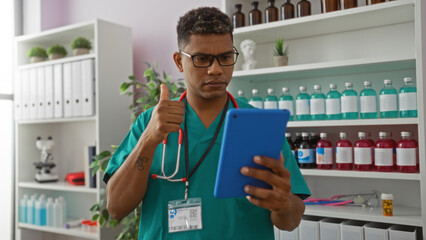 Young hispanic man in scrubs with glasses and stethoscope gives thumbs up while looking at tablet in modern clinic workplace