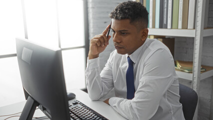 Confident hispanic man working diligently on a computer in a modern office setting with a thoughtful expression