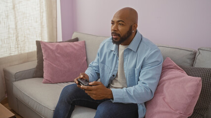 Young man playing video games in a cozy living room setting at home with a beard and bald head, sitting on a sofa amid pillows in an apartment interior