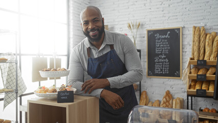 Handsome young man with a beard standing confidently in a bakery wearing an apron, surrounded by...