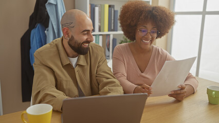 A multicultural couple laugh while reviewing a document together in a modern home office setting.