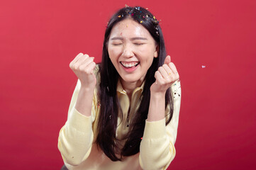 Asian woman smiling with hands raised, covered in colorful confetti, wearing a yellow sweater,...