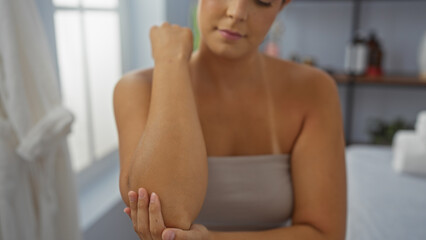 Fototapeta premium Young hispanic woman in a spa room gently touching her elbow while wearing a towel indoors