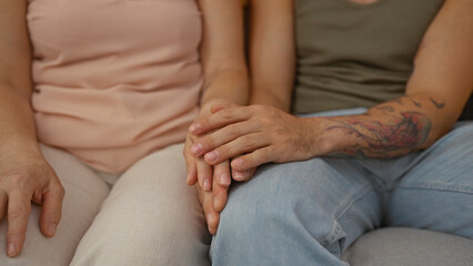 Mother and son holding hands together on a couch inside a living room, showcasing family love and strong relationship in an indoor home setting with an adult woman and man