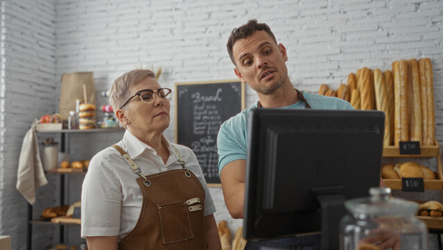 Woman and man, bakery workers, discussing at a computer in a cozy indoor shop with shelves of bread and pastries visible in the background