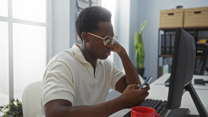 Handsome young man in an office setting, focused on his smartphone by his desk, with a computer and office supplies in the background