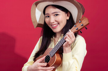 Asian woman in a yellow sweater, wearing a large, floppy straw hat, holding a ukulele and smiling off to the side. She stands against a solid red background, giving a cheerful and casual vibe.