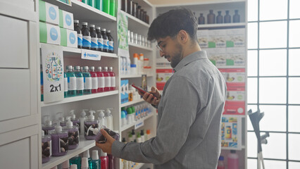 A bearded young man uses a smartphone to photograph products at a well-stocked indoor pharmacy.